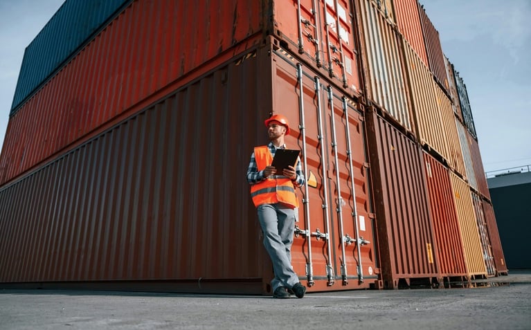 man standing in front of stacked shipping containers holding a checklist outside in spring